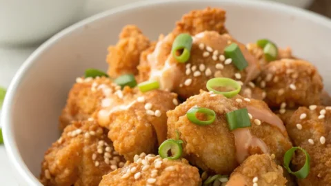 Crispy Bang Bang Chicken coated in creamy sweet-spicy sauce with green onions and sesame seeds in a white bowl.