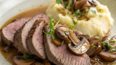 Sliced tender beef tongue served on a rustic ceramic plate with herbs, mushrooms, and broth.